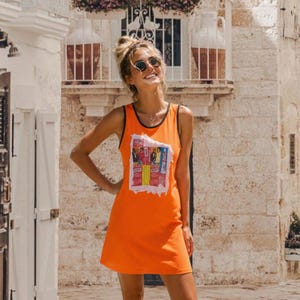 May include: A bright orange sleeveless tank dress featuring a black trim around the neckline and armholes. The center of the dress displays a rectangular graphic print with a distressed white border, containing four distinct colorful panels.