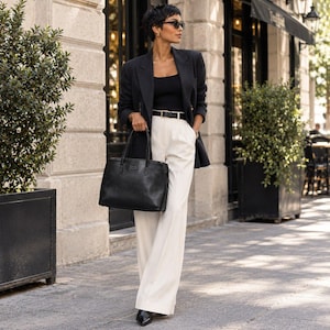 Pode incluir: Um blazer preto sobre um top preto e calças brancas de cintura subida e perna larga com cinto preto. O conjunto inclui uma mala preta estruturada com duas alças, sapatos pretos de bico fino e óculos de sol escuros.