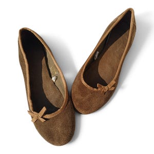 May include: A pair of brown suede ballet flats featuring a rounded toe, a low-cut topline with matching brown fabric trim, and a small decorative bow centered on the front of each shoe. The interior lining is dark brown with a textured footbed.