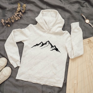 May include: A light gray hoodie with a black mountain graphic, paired with a beige pleated skirt and white sneakers. A watch and a sprig of dried leaves complete the casual outfit, set against a gray background.