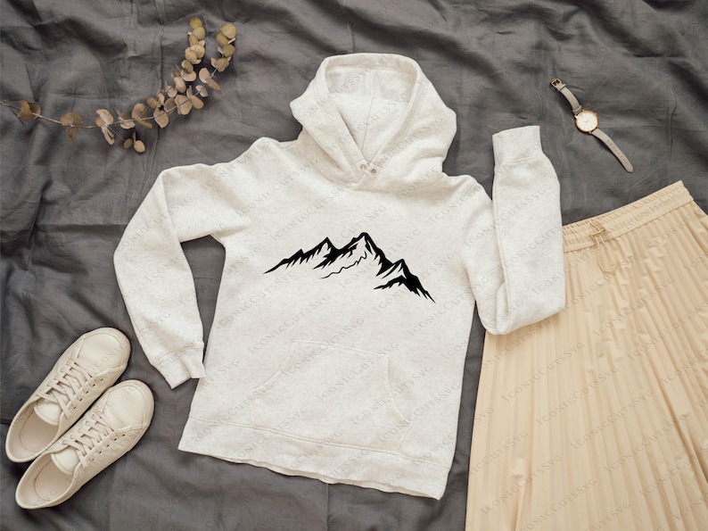 May include: A light gray hoodie with a black mountain graphic, paired with a beige pleated skirt and white sneakers. A watch and a sprig of dried leaves complete the casual outfit, set against a gray background.