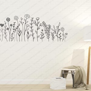 May include: A black and white line drawing of a row of stylized flowers and plants on a white wall. A white chair with a brown throw blanket is in the foreground, and a floor lamp with a white shade is to the right of the chair.