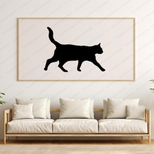 May include: A black cat silhouette in a walking pose is printed on a white background in a framed picture hanging on a white wall. A white sofa with cushions is in the foreground.