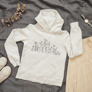 May include: A white hoodie with a black line drawing of flowers on the front. The hoodie has a drawstring hood and a kangaroo pocket.