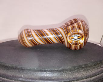 May include: A glass pipe with a rounded bowl and a long stem. The pipe features a pattern of brown, white, and orange stripes. The bowl has a clear, circular design with a logo. The pipe is resting on a black surface.