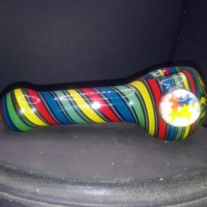 May include: A colorful glass pipe with a striped pattern in red, yellow, green, and blue. The pipe features a round bowl with a design of colorful puzzle pieces. The pipe is resting on a dark surface.