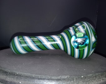 May include: Glass pipe with green, white, and blue striped pattern. The pipe has a clear, round bowl with a blue and white emblem. The body of the pipe is cylindrical with a curved mouthpiece.