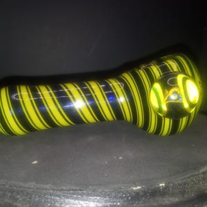 May include: A glass pipe with a yellow and black striped pattern. The pipe has a clear, rounded bowl and a curved stem. The design is a spiral of yellow and black stripes. The pipe is resting on a dark surface.