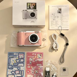 May include: A pink digital camera with a 4K lens, a white box labelled LK-007, a user manual, a white USB cable, a grey wrist strap, two sheets of character stickers, a black USB card reader, and a small memory card in a clear plastic case.
