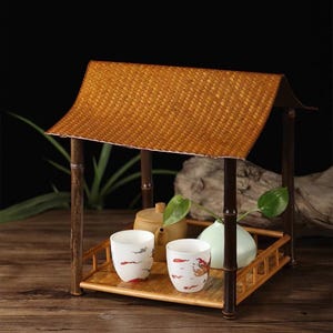 Peut inclure: Présentoir à thé en bambou avec un toit tressé et un plateau. Le plateau contient deux tasses à thé blanches avec des accents rouges, une théière et un petit vase avec des feuilles vertes. L'ensemble est posé sur une surface en bois.