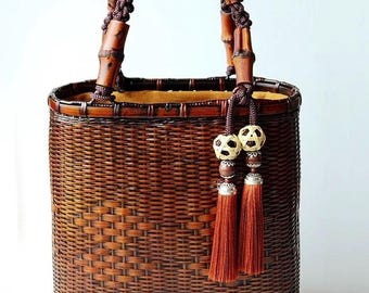 May include: A brown woven bamboo tote bag with a brown rope handle and two brown tassels. The bag has a light brown fabric lining and two decorative brown and white balls.