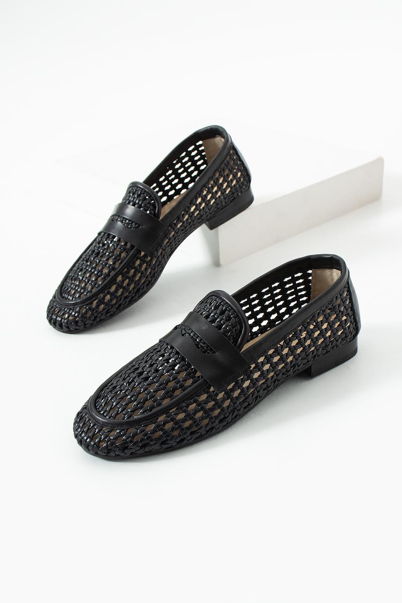 May include: A pair of black woven leather loafers with a classic penny strap design. The shoes have a low heel and a breathable, open weave pattern. The loafers are displayed on a white surface.