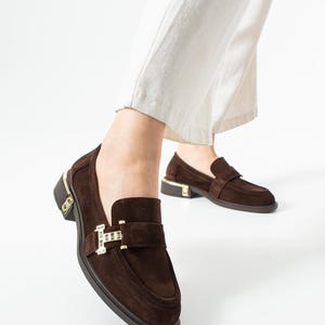Puede incluir: Mocasines de ante marr&oacute;n con detalles dorados. Los zapatos presentan un dise&ntilde;o cl&aacute;sico sin cordones con una correa en la parte superior y un tac&oacute;n peque&ntilde;o. Los detalles dorados son visibles en el tal&oacute;n y la hebilla de la correa.