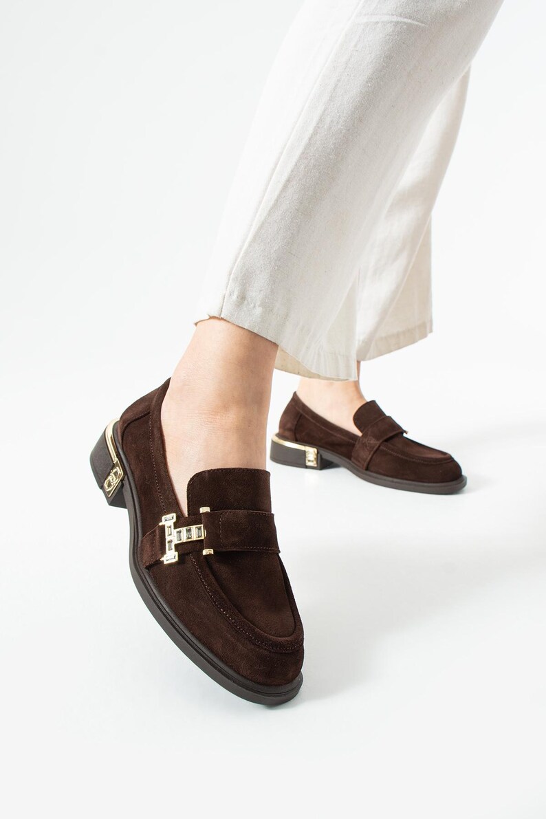 Puede incluir: Mocasines de ante marr&oacute;n con detalles dorados. Los zapatos presentan un dise&ntilde;o cl&aacute;sico sin cordones con una correa en la parte superior y un tac&oacute;n peque&ntilde;o. Los detalles dorados son visibles en el tal&oacute;n y la hebilla de la correa.