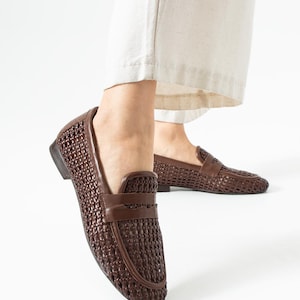 May include: Brown woven leather loafers. These slip-on shoes feature a textured, basket-weave design and a smooth leather strap across the top. The shoes are a dark brown color and have a low heel. The shoes are shown with cream-colored pants.