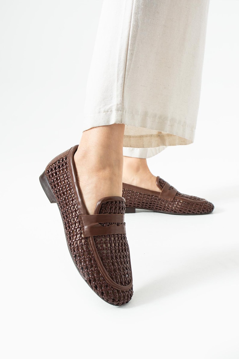 May include: Brown woven leather loafers. These slip-on shoes feature a textured, basket-weave design and a smooth leather strap across the top. The shoes are a dark brown color and have a low heel. The shoes are shown with cream-colored pants.