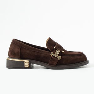 Puede incluir: Mocasines de ante marr&oacute;n con detalles dorados. Estos zapatos tienen una puntera cuadrada, un tac&oacute;n bajo y una hebilla decorativa. El tal&oacute;n tiene un ribete dorado y un logotipo. Altura del tac&oacute;n: aproximadamente 2,5 cm.