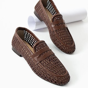 May include: A pair of brown woven leather loafers. The shoes feature a woven design on the upper and a leather strap across the vamp. The soles are a darker brown, and the shoes are set against a white background.