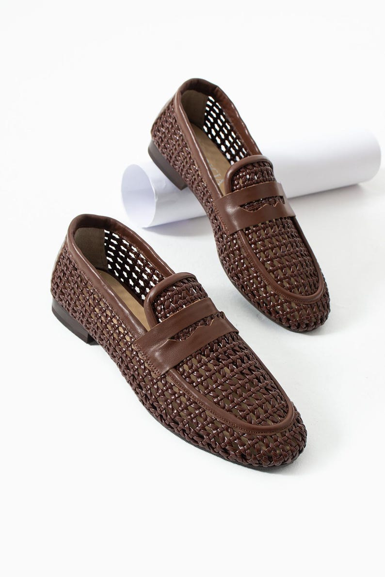 May include: A pair of brown woven leather loafers. The shoes feature a woven design on the upper and a leather strap across the vamp. The soles are a darker brown, and the shoes are set against a white background.