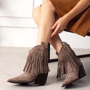 Puede incluir: Botines de ante color topo con punta afilada, tacón de bloque apilado en marrón oscuro y una capa de flecos decorativos alrededor de la caña. Cada bota incluye una pequeña lengüeta vertical en el lateral asegurada con un pequeño remache metálico dorado.