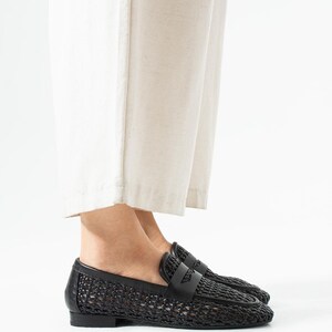 May include: Black woven leather loafers with a classic penny loafer design. The shoes feature a woven pattern on the vamp and sides, with a smooth leather strap across the top. The soles are flat and the shoes are shown from a side view.