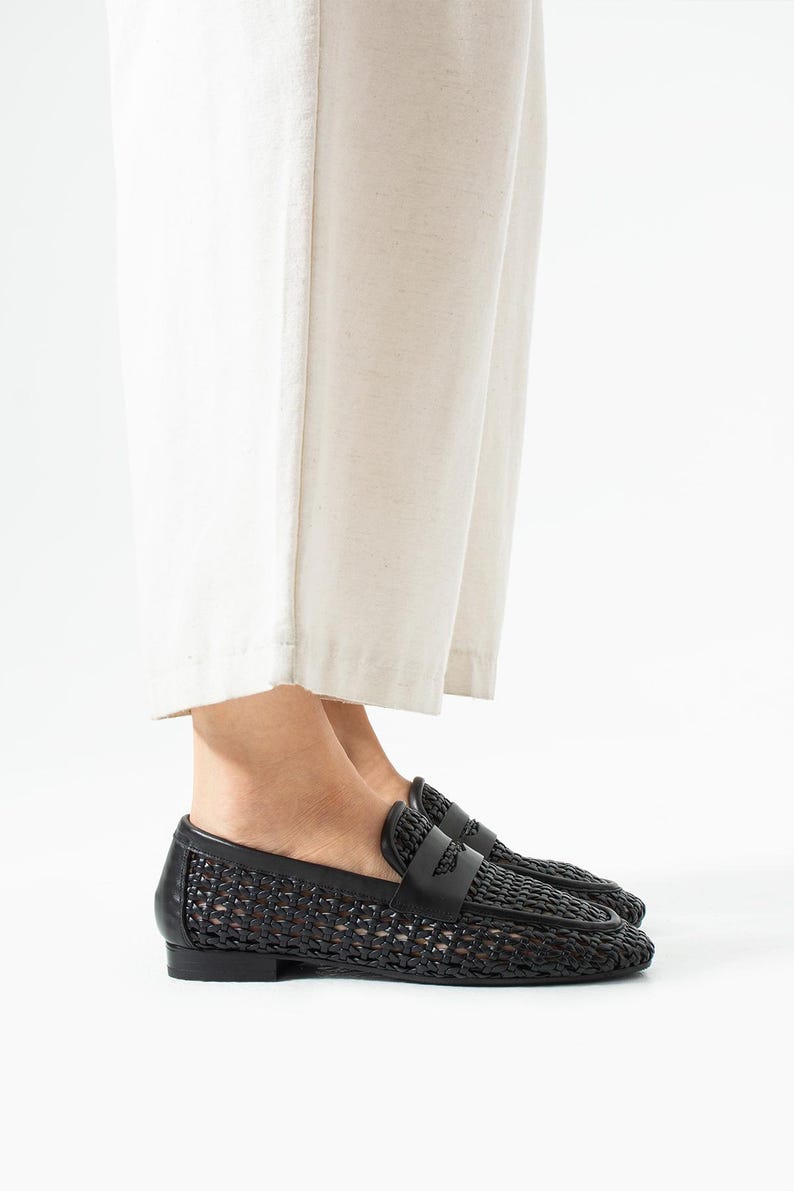 May include: Black woven leather loafers with a classic penny loafer design. The shoes feature a woven pattern on the vamp and sides, with a smooth leather strap across the top. The soles are flat and the shoes are shown from a side view.