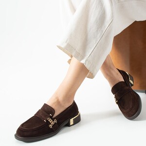 Puede incluir: Mocasines de ante marr&oacute;n con detalles dorados. Estos zapatos tienen un dise&ntilde;o cl&aacute;sico con punta redonda y tac&oacute;n bajo. Los detalles dorados est&aacute;n en el tal&oacute;n y la correa. Los zapatos se usan con pantalones blancos.