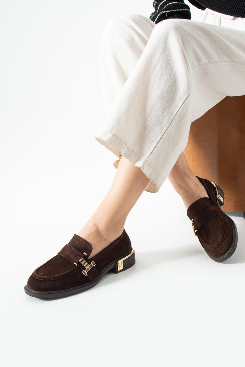 Puede incluir: Mocasines de ante marr&oacute;n con detalles dorados. Estos zapatos tienen un dise&ntilde;o cl&aacute;sico con punta redonda y tac&oacute;n bajo. Los detalles dorados est&aacute;n en el tal&oacute;n y la correa. Los zapatos se usan con pantalones blancos.