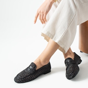 May include: Black woven leather loafers with a classic penny loafer design. The shoes have a textured, open weave pattern and a smooth leather strap across the top. The shoes are shown on a person wearing cream-colored pants.