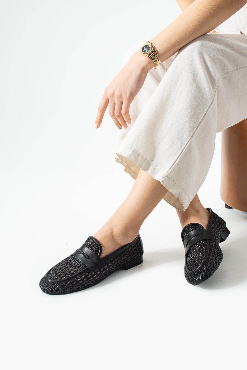 May include: Black woven leather loafers with a classic penny loafer design. The shoes have a textured, open weave pattern and a smooth leather strap across the top. The shoes are shown on a person wearing cream-colored pants.