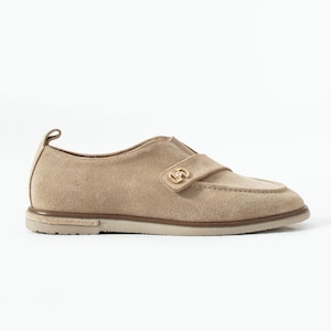 Puede incluir: Zapato tipo mocasín de ante color beige con una correa en la parte superior con una hebilla dorada. El zapato tiene una puntera redondeada y una suela clara con ribete marrón. El talón tiene un pequeño lazo.