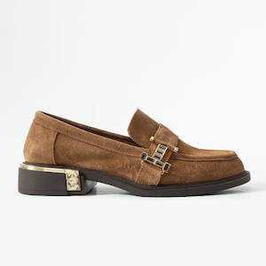 Puede incluir: Mocasín de ante marrón con hebilla y ribete dorados. El zapato tiene un tacón bajo y un diseño clásico. El mocasín es un modelo sin cordones, perfecto para uso informal o semiformal.