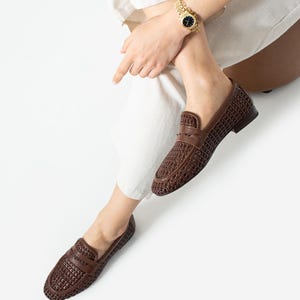 May include: Brown woven leather loafers with a classic design. The shoes have a textured pattern and a low heel. A gold watch with a black face is visible on the wrist. The person is wearing white pants.