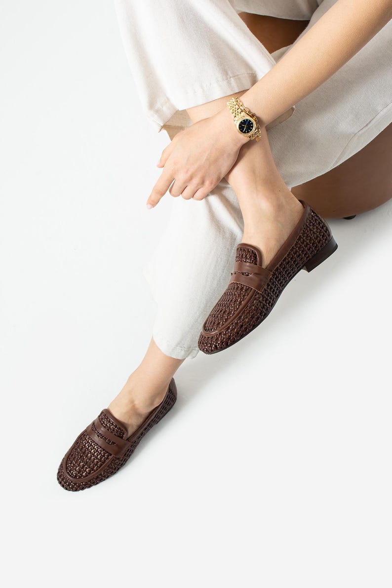 May include: Brown woven leather loafers with a classic design. The shoes have a textured pattern and a low heel. A gold watch with a black face is visible on the wrist. The person is wearing white pants.