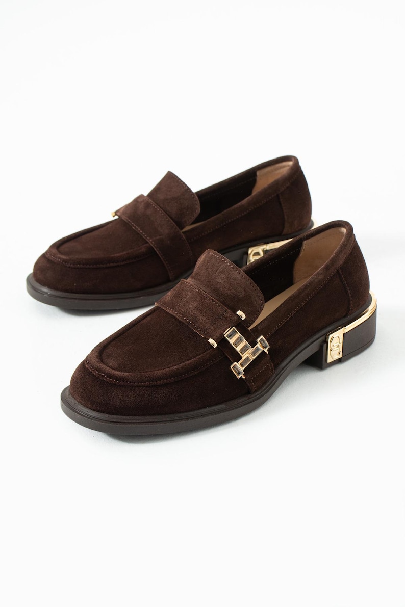 Puede incluir: Un par de mocasines de ante marr&oacute;n con hebilla y detalles de tac&oacute;n dorados. Los zapatos tienen un dise&ntilde;o cl&aacute;sico sin cordones con punta redondeada y tac&oacute;n bajo. Los mocasines se muestran sobre un fondo blanco.