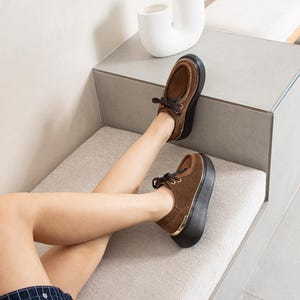 May include: A pair of brown suede shoes with dark brown laces and a thick black platform sole. The shoes feature a rounded toe, gold-tone metal eyelets for the laces, and a metallic gold accent strip integrated into the heel of the black platform base.