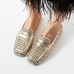 Puede incluir: Un par de mocasines dorados con una parte superior de malla tejida, una tira de cuero metalizado sobre el empeine y una puntera cuadrada. Los zapatos se llevan con una falda negra con un dobladillo de flecos.