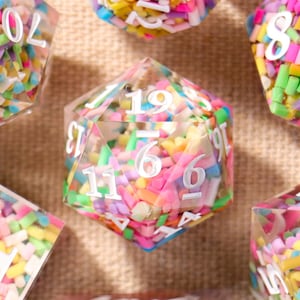 May include: Set of seven polyhedral dice with clear resin bodies filled with colorful sprinkles. Each die features white numbers. The dice are in various geometric shapes, including a d4, d6, d8, d10, d12, and d20, perfect for tabletop gaming.