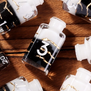 May include: Seven decorative glass bottles with white tops and black and white bodies. Each bottle has gold accents and white numbers. The bottles are arranged on a wooden surface.