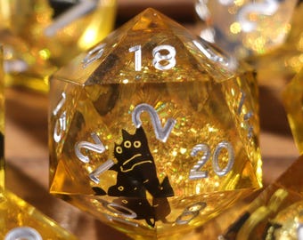 May include: A set of polyhedral dice with a golden, glittery interior and black cat illustrations. The dice have silver numbering and are made of a transparent, amber-colored resin. The dice are designed for tabletop gaming.
