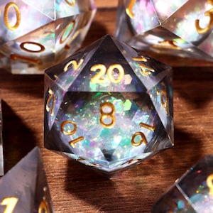 May include: Set of clear, multi-sided dice with gold numbering and iridescent glitter inclusions. The dice are designed for tabletop games and are displayed on a wooden surface.