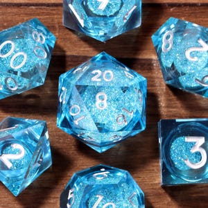 Op de afbeelding: Set van zeven helderblauwe polyhedrale dobbelstenen met zilveren cijfers, ontworpen voor tabletop-rollenspellen. De dobbelstenen zijn gerangschikt op een houten oppervlak, waarbij hun veelzijdige vormen en interne glittereffect worden getoond. Elke dobbelsteen toont een ander nummer.