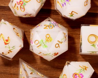 May include: Set of seven white polyhedral dice with gold numbers and floral designs in pink, yellow, and green. The dice are for role-playing games and include a d4, d6, d8, d10, d12, d20, and a cube.