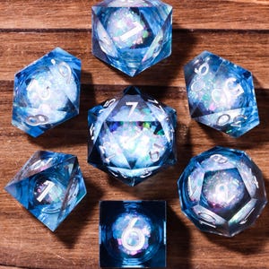 Puede incluir: Un juego de siete dados poliédricos hechos de resina azul translúcida con escamas de purpurina iridiscente incrustadas. Cada dado presenta bordes afilados y números blancos claros. La colección incluye varias formas como d20, d12, d10, d8, d6 y d4.