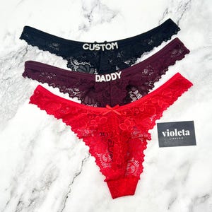 May include: Three lace thongs in black, dark burgundy, and bright red. The black pair features the word CUSTOM in silver rhinestones, the burgundy pair features the word DADDY in silver rhinestones, and the red pair has a small centre bow.