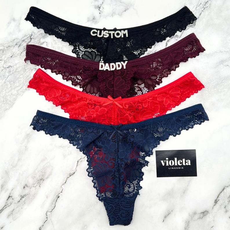 May include: Four lace thongs in black, burgundy, red, and navy blue. The black pair features the word CUSTOM in silver rhinestones, the burgundy pair features DADDY in silver rhinestones, and all pairs include a small centre bow. A black tag reads violeta LINGERIE.