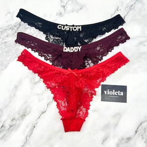 May include: Three pairs of lace thong underwear in black, burgundy, and red. The black pair features the text CUSTOM in rhinestones, the burgundy pair features the text DADDY in rhinestones, and the red pair has a small bow. A card reads violeta LINGERIE.