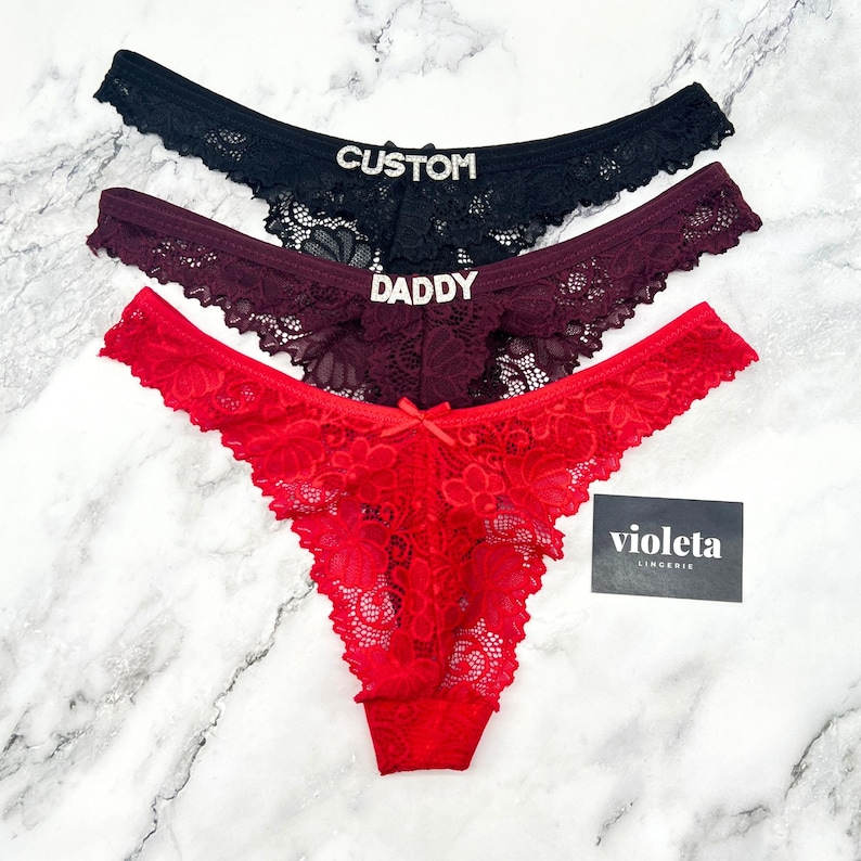 May include: Three pairs of lace thong underwear in black, burgundy, and red. The black pair features the text CUSTOM in rhinestones, the burgundy pair features the text DADDY in rhinestones, and the red pair has a small bow. A card reads violeta LINGERIE.