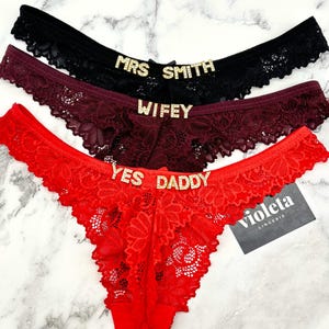 May include: Three pairs of lacy underwear with gold glitter lettering. The top pair is black with "MRS SMITH" written on it. The middle pair is burgundy with "WIFEY" written on it. The bottom pair is red with "YES DADDY" written on it.