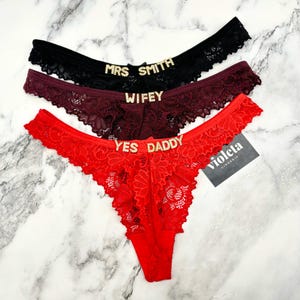 May include: Three pairs of lace thong underwear in black, burgundy, and red. Each pair features gold-toned rhinestone lettering on the front waistband. The black pair reads MRS SMITH, the burgundy pair reads WIFEY, and the red pair reads YES DADDY.